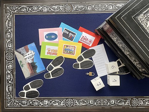 Verschieden farbige Kärtchen, drei Würfel und kleine Schuhabdrücke liegen neben einer Schatulle auf einem blauen Tisch.